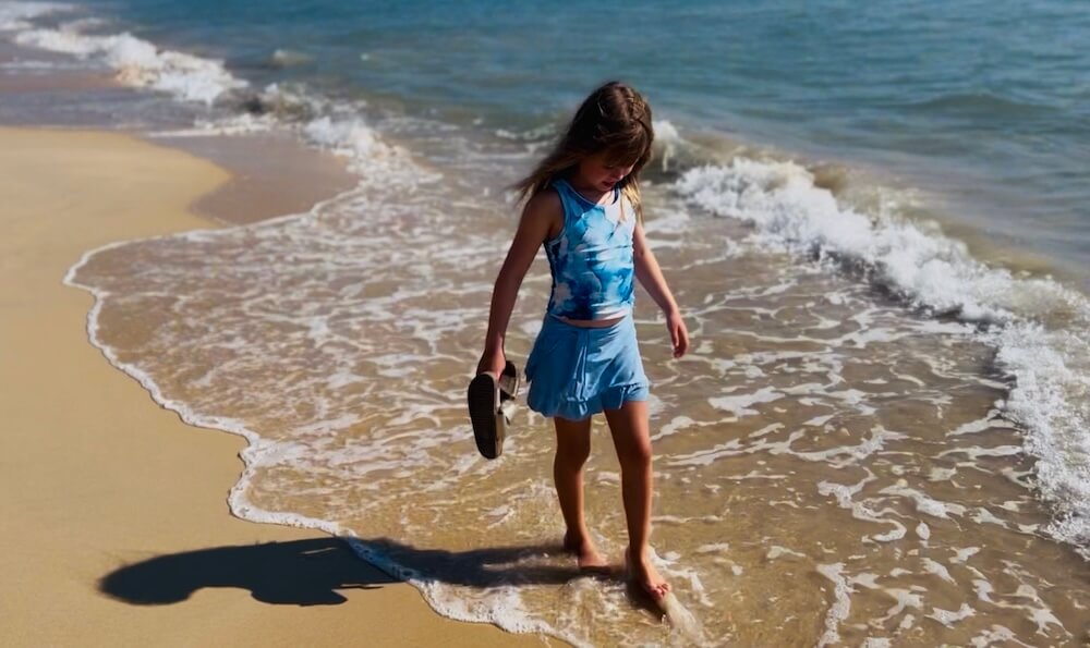Solitude at Bor Dan A young girl in a blue swimsuit walks along the shoreline of Bor Dan Beach, carrying her sandals as the gentle tide washes over her feet.