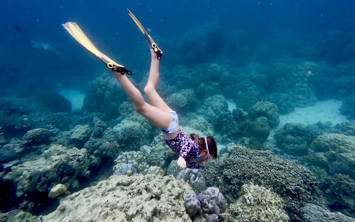 A young girl with yellow fins diving down toward large coral formations in the clear blue water of the Surin Islands.