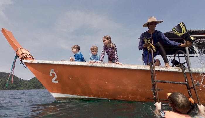 Three young children lean over the side of an orange longtail boat while a guide prepares fins and another child swims in the water below.