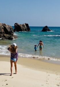 A woman and child wade into the turquoise water of a hidden cove surrounded by large rock formations, while a young girl walks on the sand in the foreground.