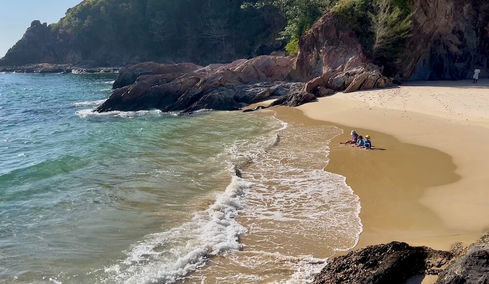 A Desert Island Discovery Three children sit on the edge of the tide on a golden sand beach, framed by dramatic rocky cliffs and a jungle-covered escarpment.