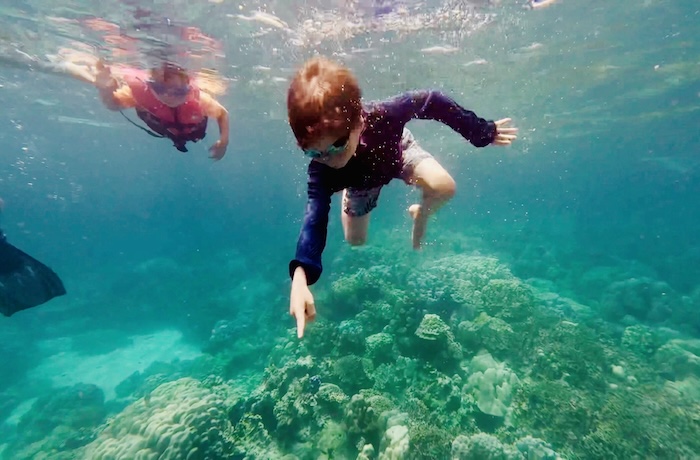 An underwater shot of a young boy in a swim shirt and goggles pointing at a coral reef while another child in a life jacket swims nearby in the Surin Islands.