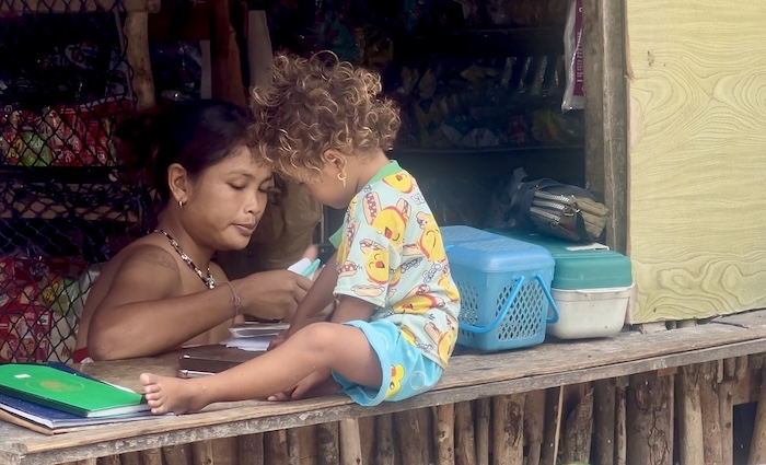 A Moken woman and a young child with curly hair sitting together at a wooden counter in their village on the Surin Islands.