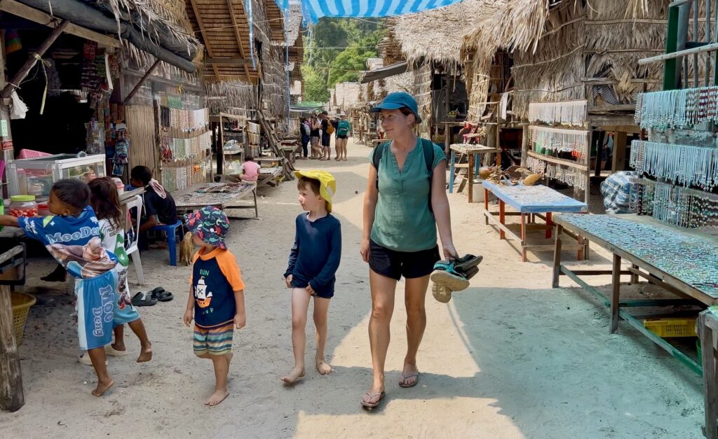 A woman and two young children walk down the central sandy path of the Moken Village on the Surin Islands, flanked by stilted bamboo houses and souvenir tables.