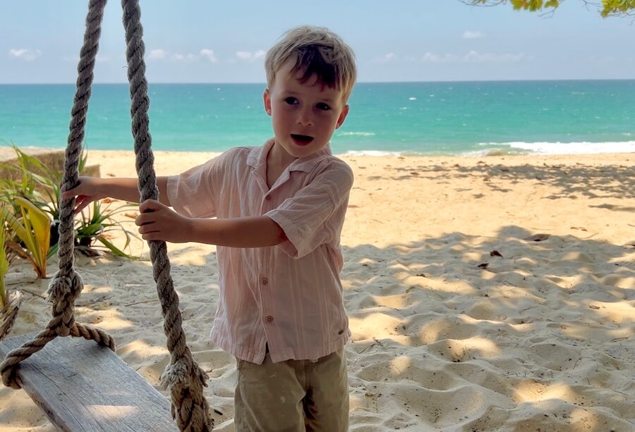 Waiting Out the Heat A young boy stands next to a wooden rope swing in the shade of a large tree on a sandy beach, with the turquoise Andaman Sea in the background.