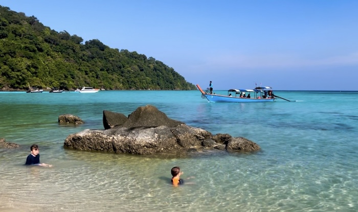 Two young boys playing in the shallow, clear turquoise water of a quiet beach in the Surin Islands as a longtail boat passes in the background.