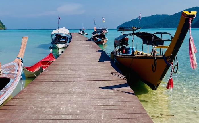 A wooden pier stretching out into clear turquoise water with several colorful longtail boats moored on either side in the Surin Islands.