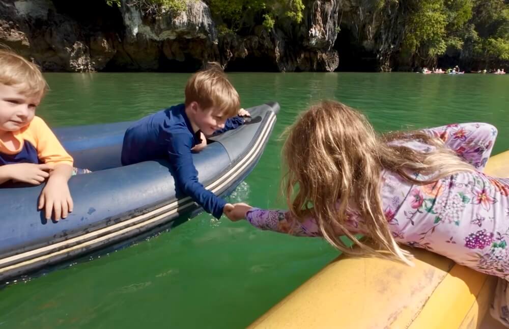 Three children leaning out of inflatable canoes to hold hands over bright green water with limestone cliffs and sea caves in the background.