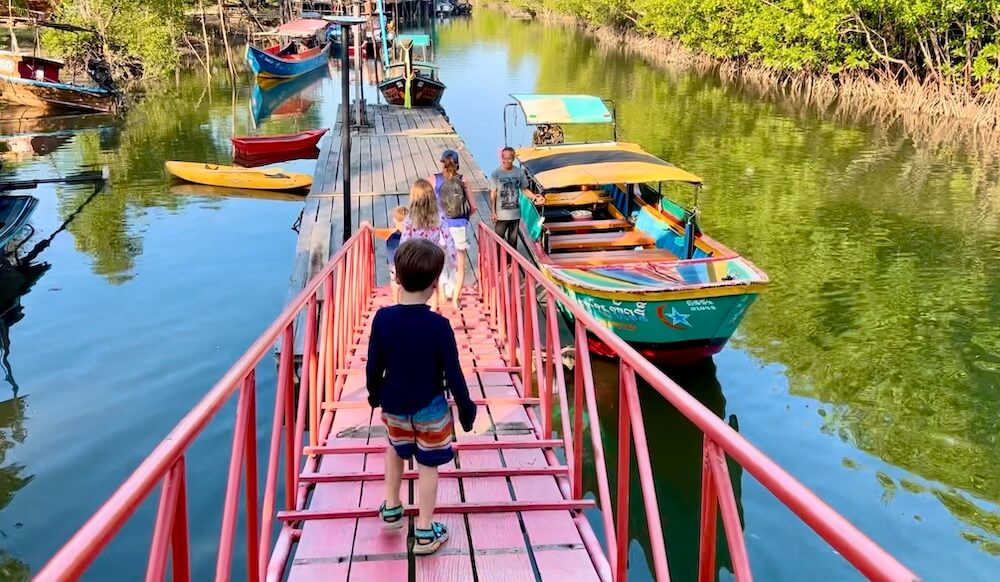 A family walking down a red metal ramp onto a wooden pier to board a colorful Thai longtail boat in a mangrove channel.