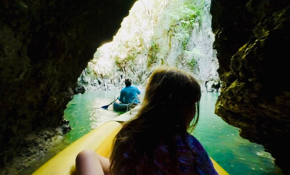 An inflatable kayak emerging from a dark limestone cave tunnel into a bright, green water lagoon surrounded by sheer cliffs at Panak Island.