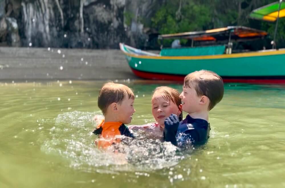 Three children splashing and swimming in the shallow green water of Phang Nga Bay, Thailand, with a colorful longtail boat in the background.