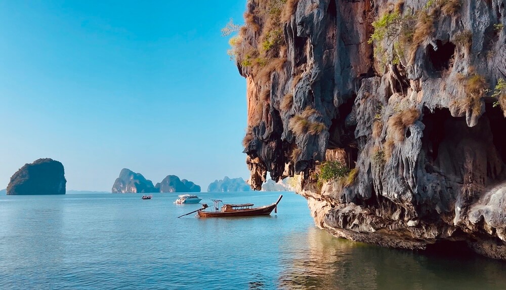 A wooden Thai longtail boat anchored next to a sheer limestone cliff featuring natural openings and windows in Phang Nga Bay, Thailand.