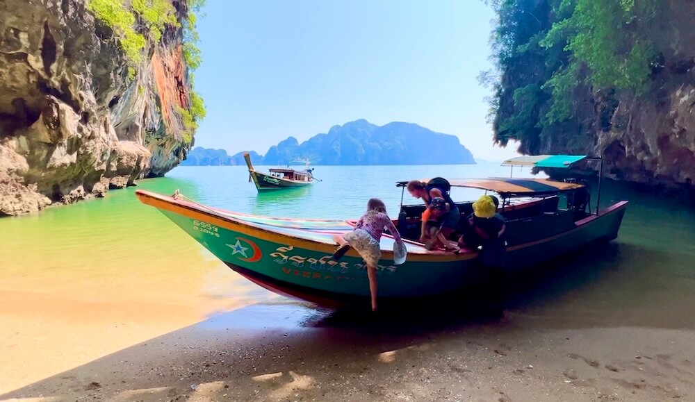 A family stepping off a colorful Thai longtail boat onto a quiet, sandy beach nestled between towering limestone cliffs in Phang Nga Bay.
