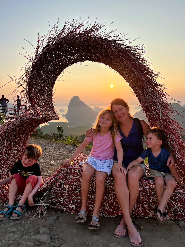 A mother and three children posing inside a large woven heart sculpture at the Samet Nangshe Viewpoint during a golden sunrise over Phang Nga Bay, Thailand.