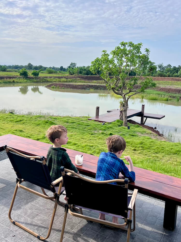 Two young boys sitting side-by-side in folding chairs at an outdoor wooden counter, eating breakfast while looking out over a peaceful lake and green lawn.