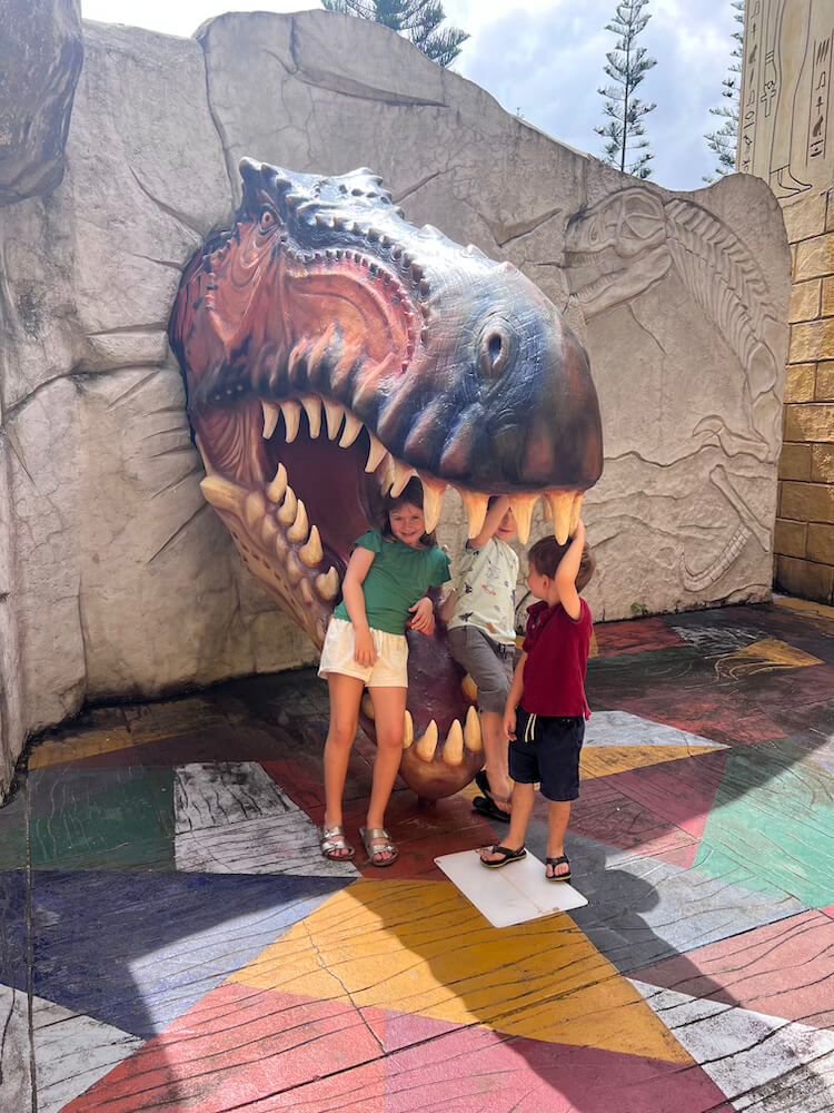 hree young children playing and posing playfully inside the open mouth of a large T-Rex dinosaur head sculpture on a colorful paved patio.
