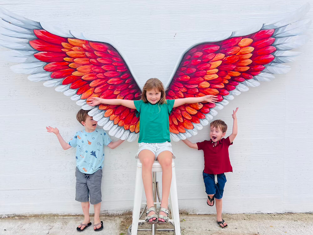 hree playful children posing in front of a white wall featuring a large, brightly painted red and orange wing mural.