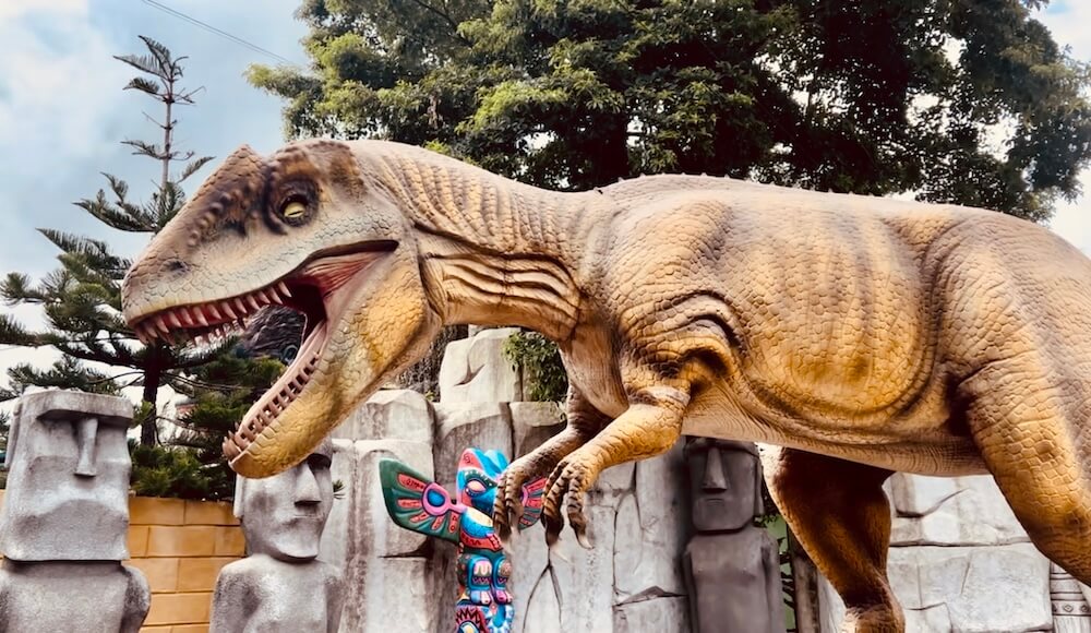 A large T-Rex dinosaur figure standing in front of Easter Island Moai statues and a colorful totem pole at Amazing Dinosaur in Khao Kho.