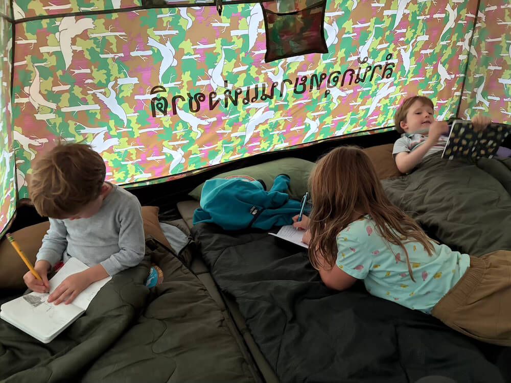Three young children lying in sleeping bags inside a colorful pattern-walled tent, writing and drawing in notebooks.