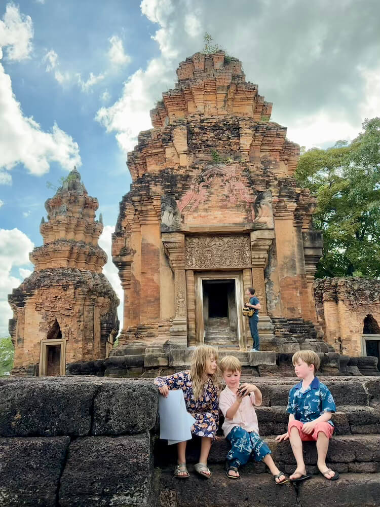 Three young children sitting on the stone steps of an ancient brick temple, with an adult standing near the temple entrance under a blue cloudy sky.