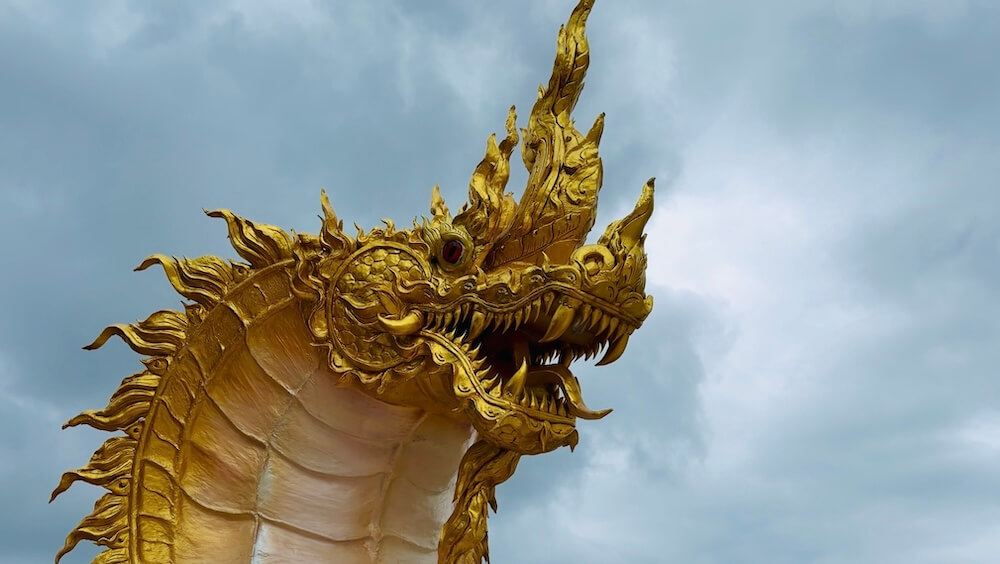 Close-up of an intricately carved golden Naga serpent head against dramatic, overcast sky at Wat Phra That Pha Sorn Kaew.