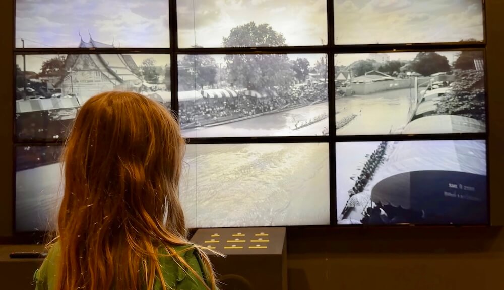 A young girl with long reddish-brown hair viewed from behind, watching a large multi-screen video display showing local boat races.