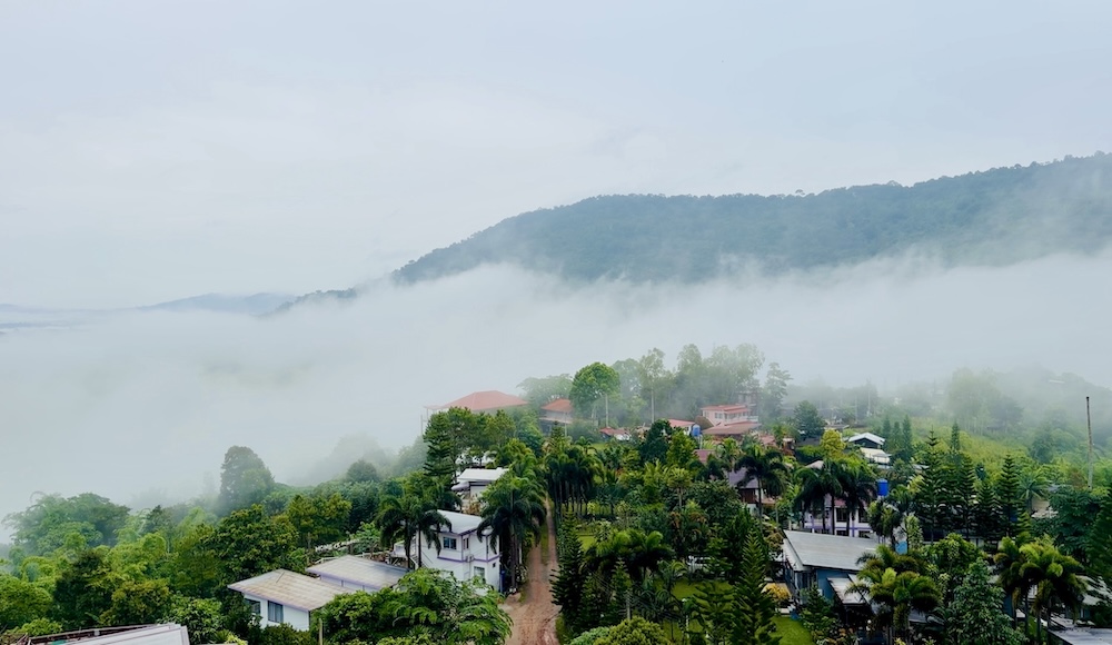 Low-hanging white clouds and mist rolling through a green valley filled with palm trees and hilltop resorts in Khao Kho, Thailand.