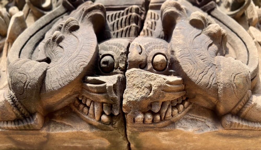 Extreme close-up of a cracked ancient Khmer sandstone lintel carving featuring a Kala face with prominent teeth at Si Thep.