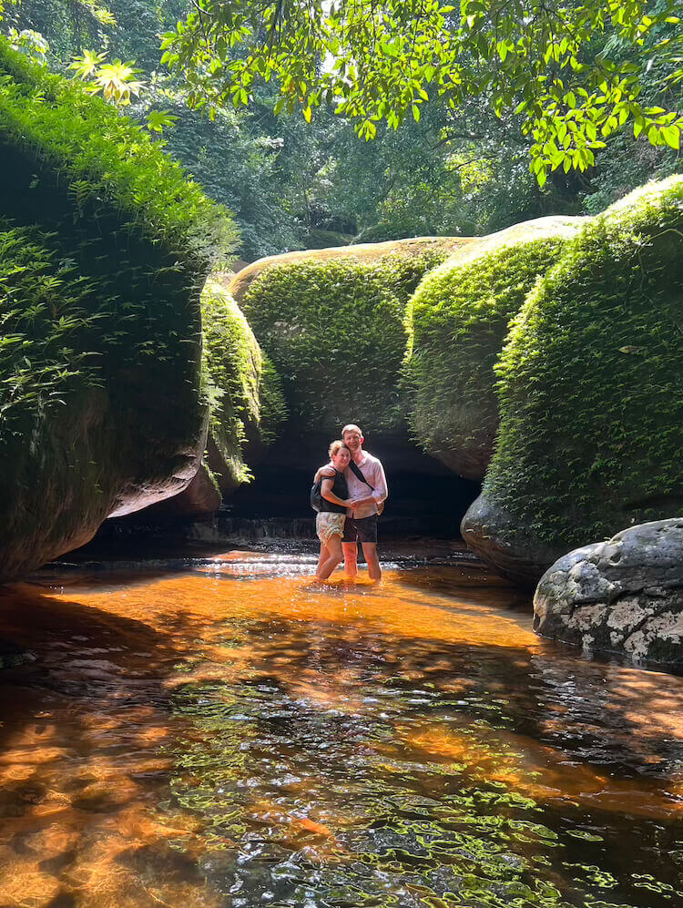 kinnaree-waterfall-thailand-jungle-trek-couple A man and woman standing together in shallow water surrounded by large mossy rock formations and dense green jungle foliage.