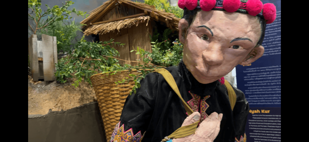 Life-size mannequin figure in traditional Nyah Kur ethnic dress carrying a woven basket in front of a rural house diorama.