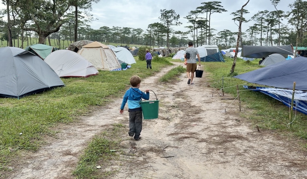 A young boy carrying a green water bucket along a dirt path through a busy tent campsite in a pine forest