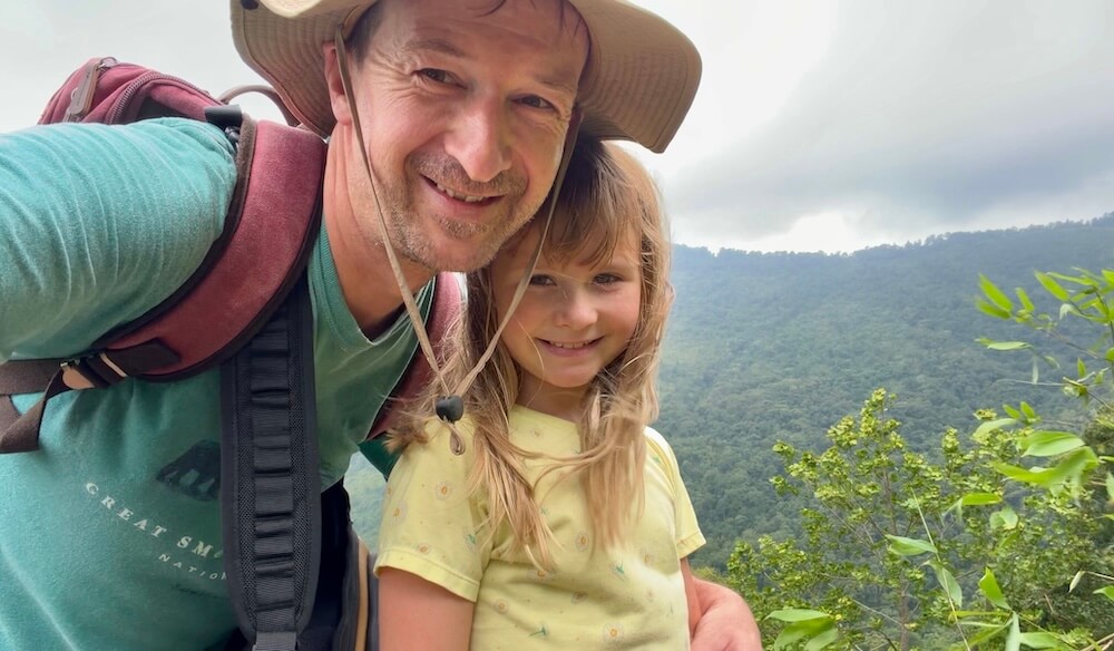 A father in a sun hat and his young daughter smiling for a selfie on a mountain ridge overlooking lush green valleys