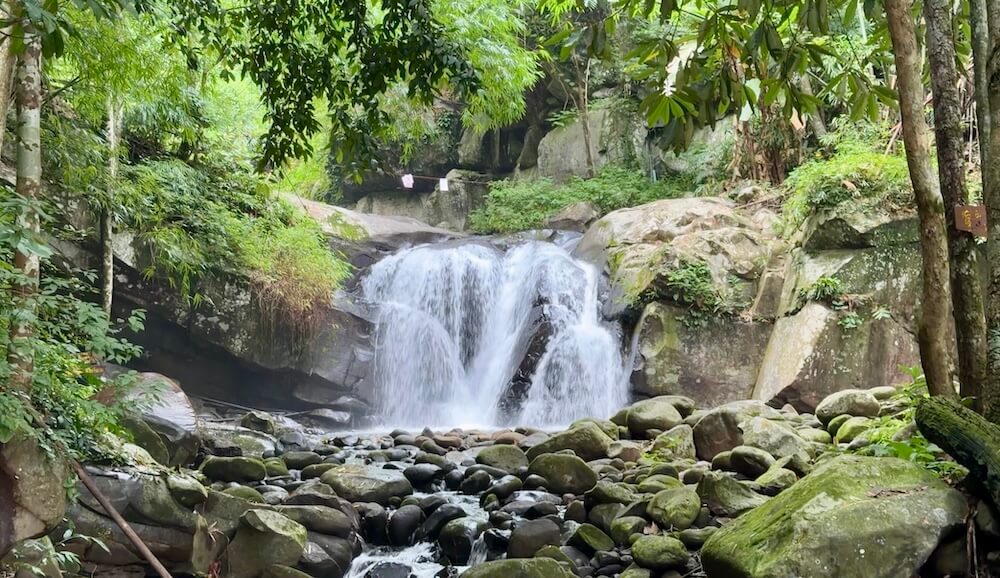 A wide forest waterfall rushing over dark rocks into a boulder-filled stream surrounded by lush green jungle foliage