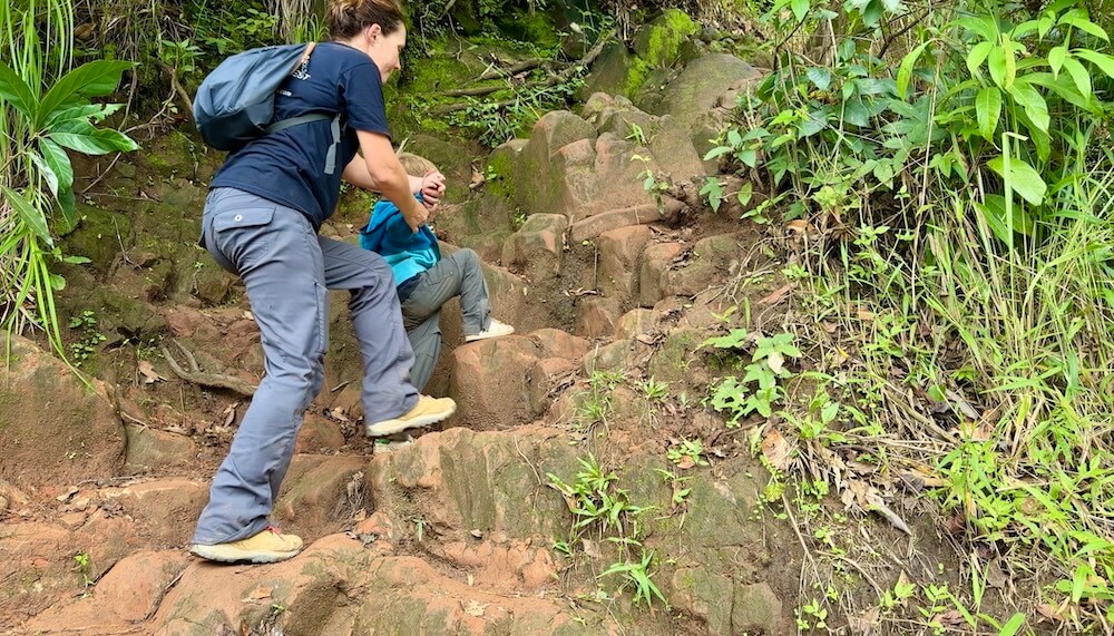 A mother guiding her young son by the hand as they step up steep, natural stone stairs on a mountain trail