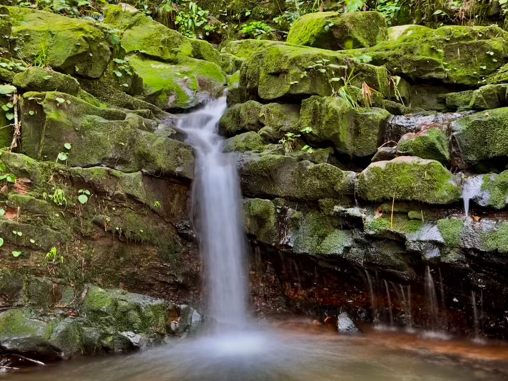 A small, picturesque waterfall cascading into a natural pool framed by dark rock walls covered in lush green moss
