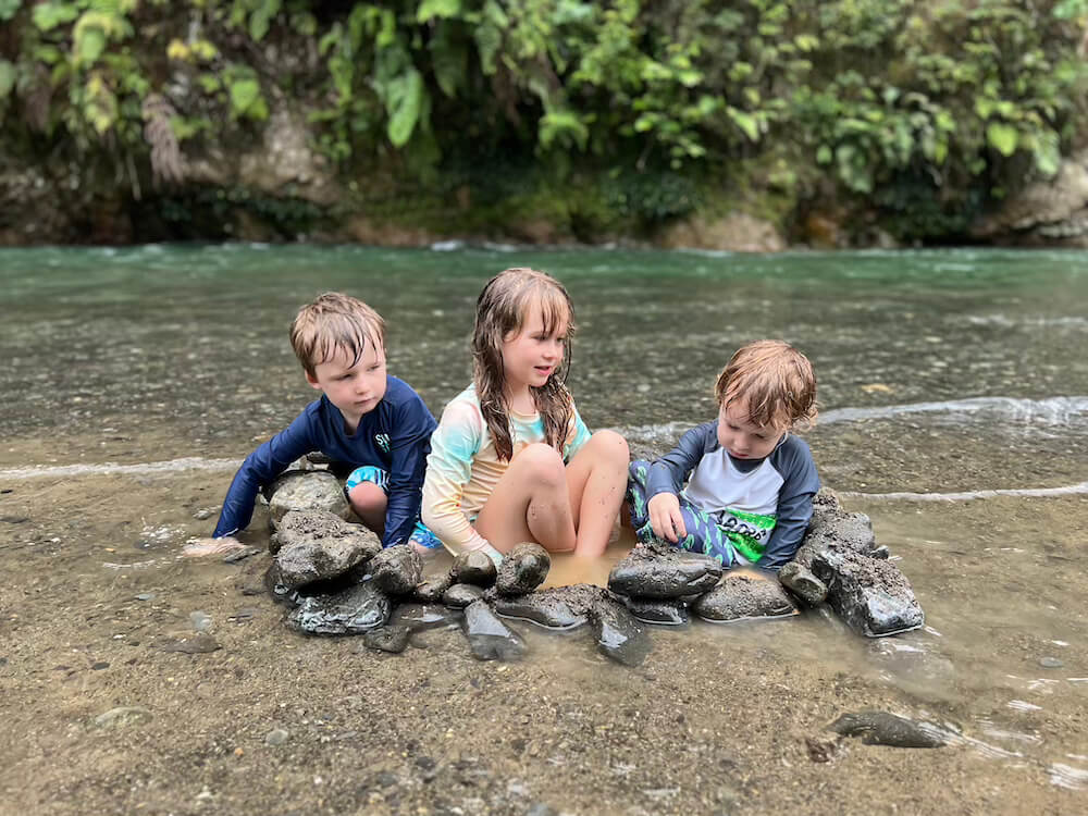 Three young children in rash guards sitting inside a small circle of rocks in a shallow river, surrounded by lush green greenery in the background.