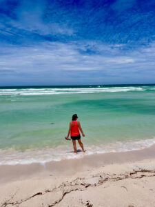 A woman in a pink top and shorts standing ankle-deep in calm turquoise ocean water along a white sandy beach under a blue sky.