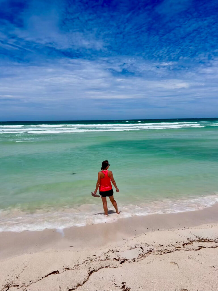A woman in a pink top and shorts standing ankle-deep in calm turquoise ocean water along a white sandy beach under a blue sky.