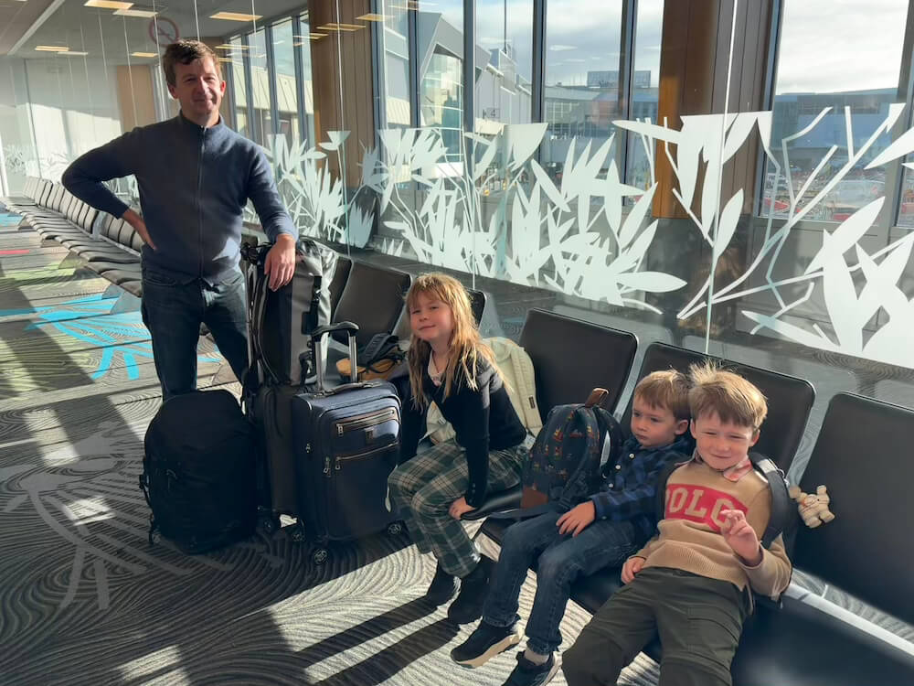 A father standing beside a stack of luggage next to his three young children sitting in terminal gate chairs at an airport.