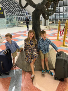 Three young children posing with their rolling suitcases around an indoor decorative tree planter in a modern airport terminal.