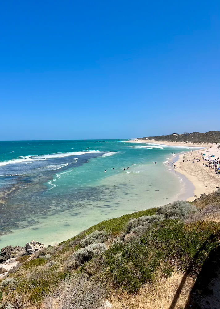 High-angle view of a long sandy beach line bordering vibrant turquoise ocean waters under a clear blue sky.