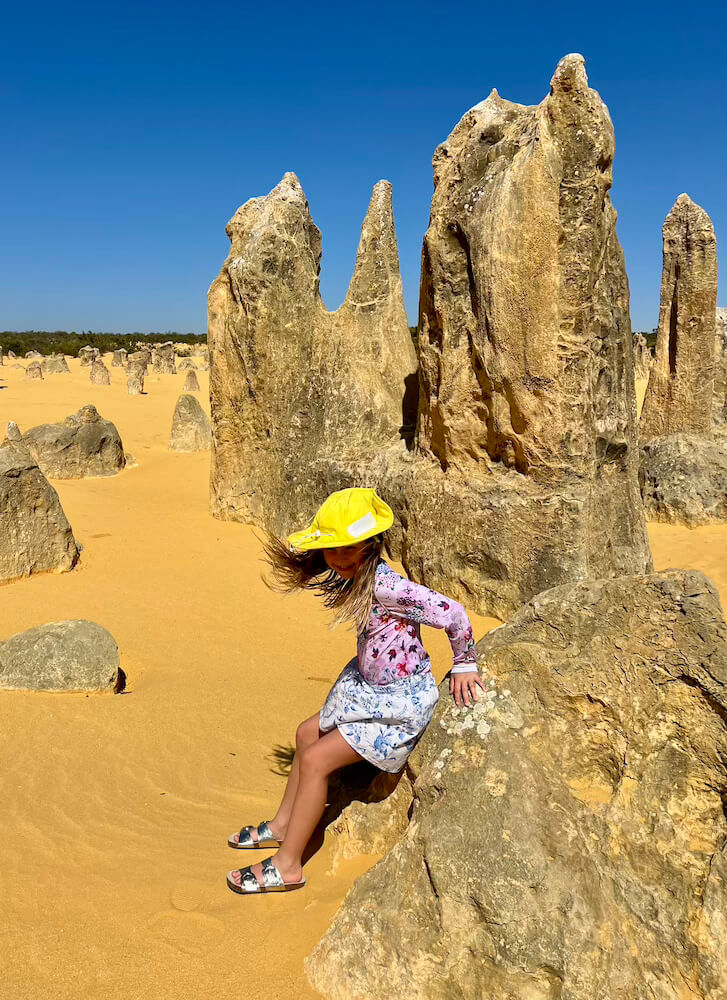 A young girl wearing a yellow sun hat sitting on a limestone rock formation in the yellow sand of the Pinnacles Desert under a bright blue sky.