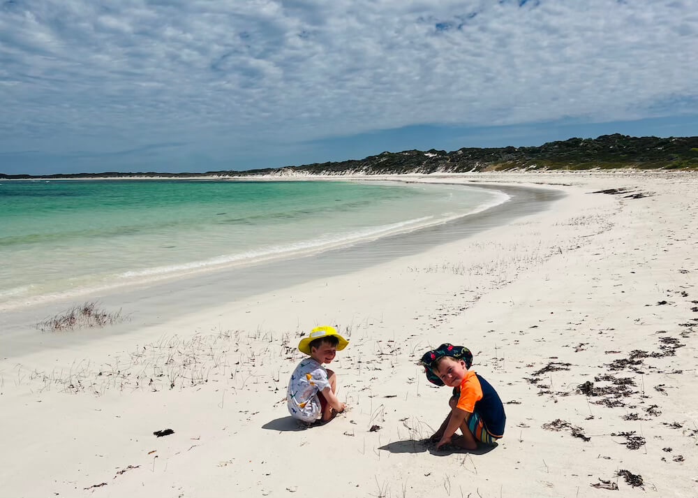 Two young boys wearing sun hats sitting and playing in the white sand of a pristine, empty beach with clear turquoise water.