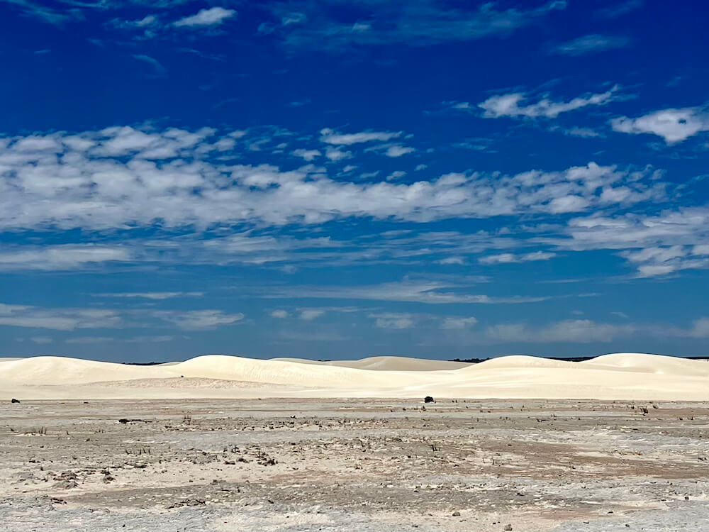 Rolling white sand dunes stretching across the horizon under a bright blue sky filled with light white clouds.