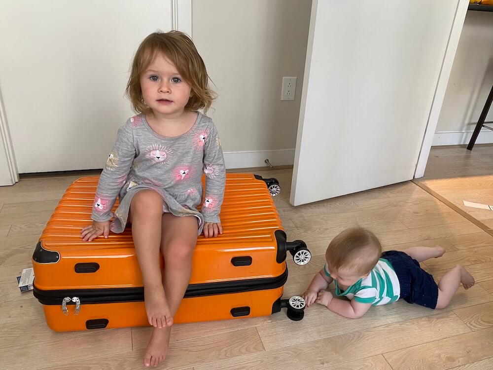 A young toddler girl sitting on top of a large orange suitcase while a baby crawls on the floor beside her.