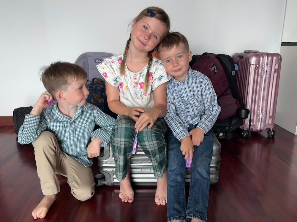 Three young siblings leaning together fondly while sitting on silver rolling luggage surrounded by backpacks and suitcases.