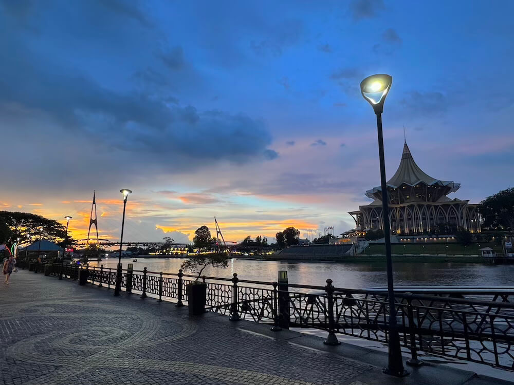 Paved waterfront promenade with street lamps along the Sarawak River at sunset, showing the Sarawak State Legislative Assembly building across the water.