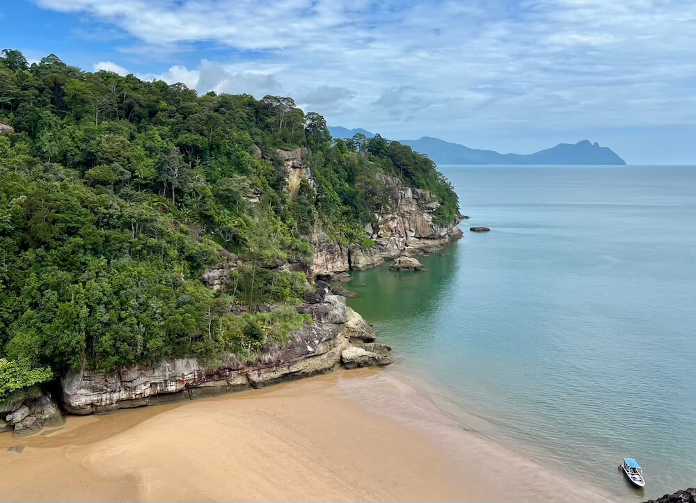 High-angle view of a sandy beach cove flanked by rainforest-covered cliffs and blue ocean at Bako National Park.