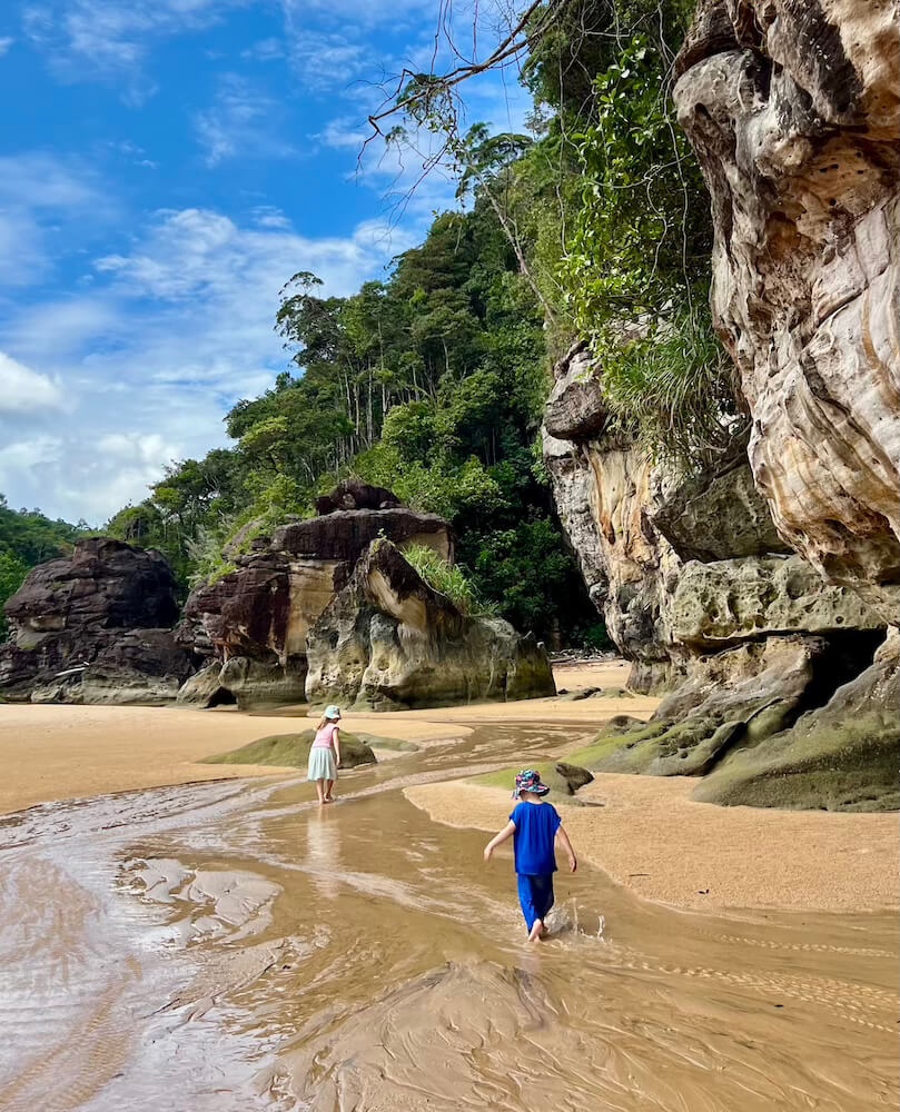 Two young children wading through a sandy stream below large sandstone cliffs at Bako National Park.