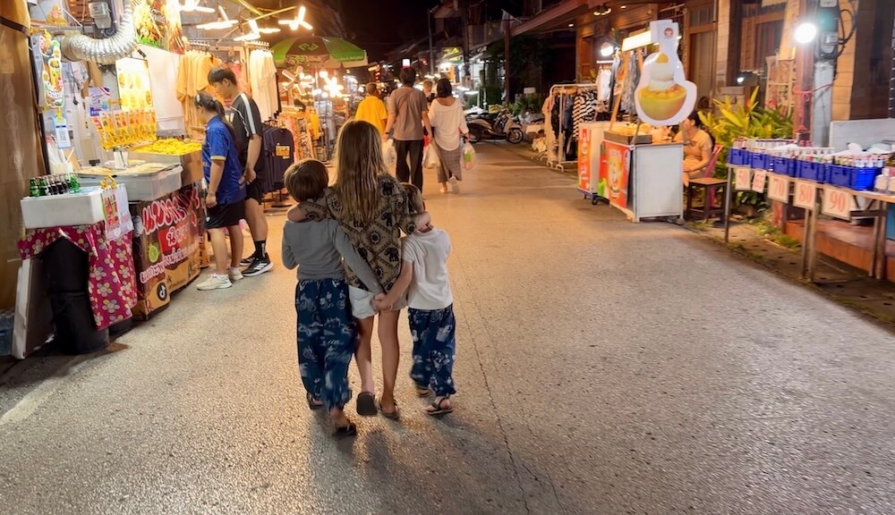 Three young siblings walking with arms wrapped around each other's shoulders down a brightly lit Thai night market street lined with food stalls.